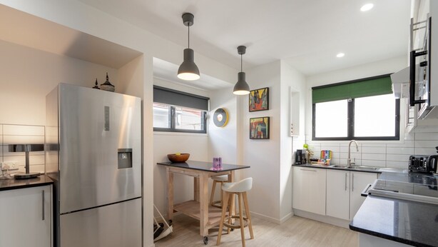 A modern kitchen with a stainless steel refrigerator, wooden bar stools, a small dining table, and pendant lighting. - Sophisticated 2-Bed Home With Alfresco Dining (Fortitude Valley)