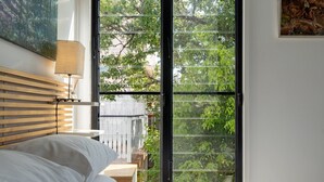 Une chambre à coucher avec un lit, une table de chevet, une lampe et une grande fenêtre donnant sur des arbres.
