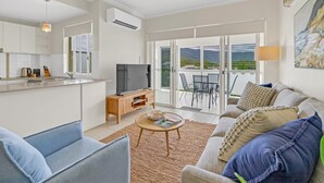 Een moderne woonkamer met een bank, salontafel en televisie. Er is een keuken met een aanrecht en apparatuur. De kamer heeft grote ramen en uitzicht op de omgeving.