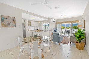 A modern living room with a glass dining table, white chairs, a grey sofa, and a large potted plant. - Trinity Beach Retreat with Pool and Scenic Views (Trinity Beach)