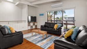 A modern living room with a black leather sofa, a wooden coffee table, and a large window with a view of palm trees. - Spacious 4-Bed Home 5-min from the Beach (Capel Sound)