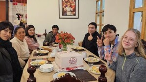 Un groupe de personnes est assis autour d’une table, devant de la nourriture et un gâteau, dans une pièce où un tableau encadré est accroché au mur.