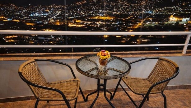 Un balcon avec des fauteuils en osier et une table en verre, d’où l’on peut admirer la vue sur la ville illuminée la nuit.