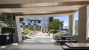 An outdoor terrace with stone flooring, wooden benches, and a view of a modern building and marina. - 1BR Apartment Near CBD with Pool (Docklands)