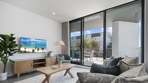A modern living room with a large TV, a wooden coffee table, a sofa, and a view of the outdoors through large sliding glass doors. - Spacious 2-Bed Apartment with Rooftop Views (North Fremantle)