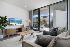 Ein modernes Wohnzimmer mit einem großen Fernseher, einem Holztisch, einer Couch und Blick ins Freie durch große Schiebeglaswände.