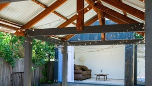 Une terrasse extérieure avec une table et des chaises en bois, des guirlandes lumineuses et une vue sur une maison en arrière-plan.