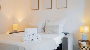 Una cama bien hecha con toallas enrolladas, dos lámparas de noche y tres cuadros en la pared.