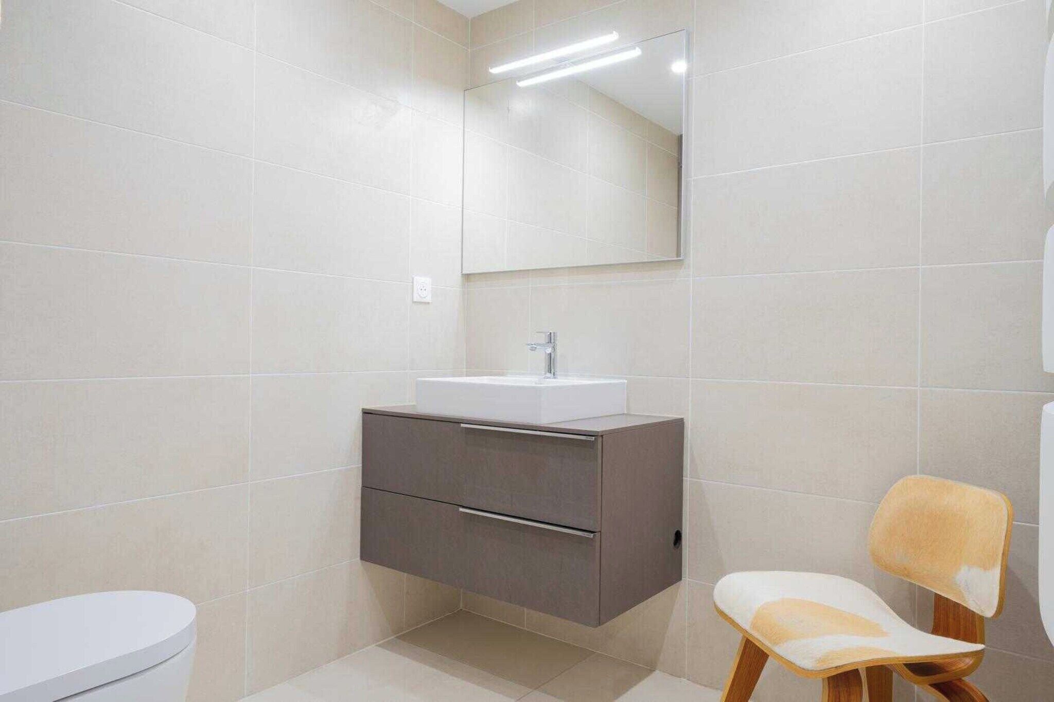 A modern bathroom with a white sink, a mirror above it, and a wooden chair.
