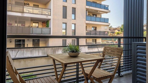 Un balcón con mesa y sillas de madera, una planta en maceta y vistas de un edificio de varios pisos con balcones.