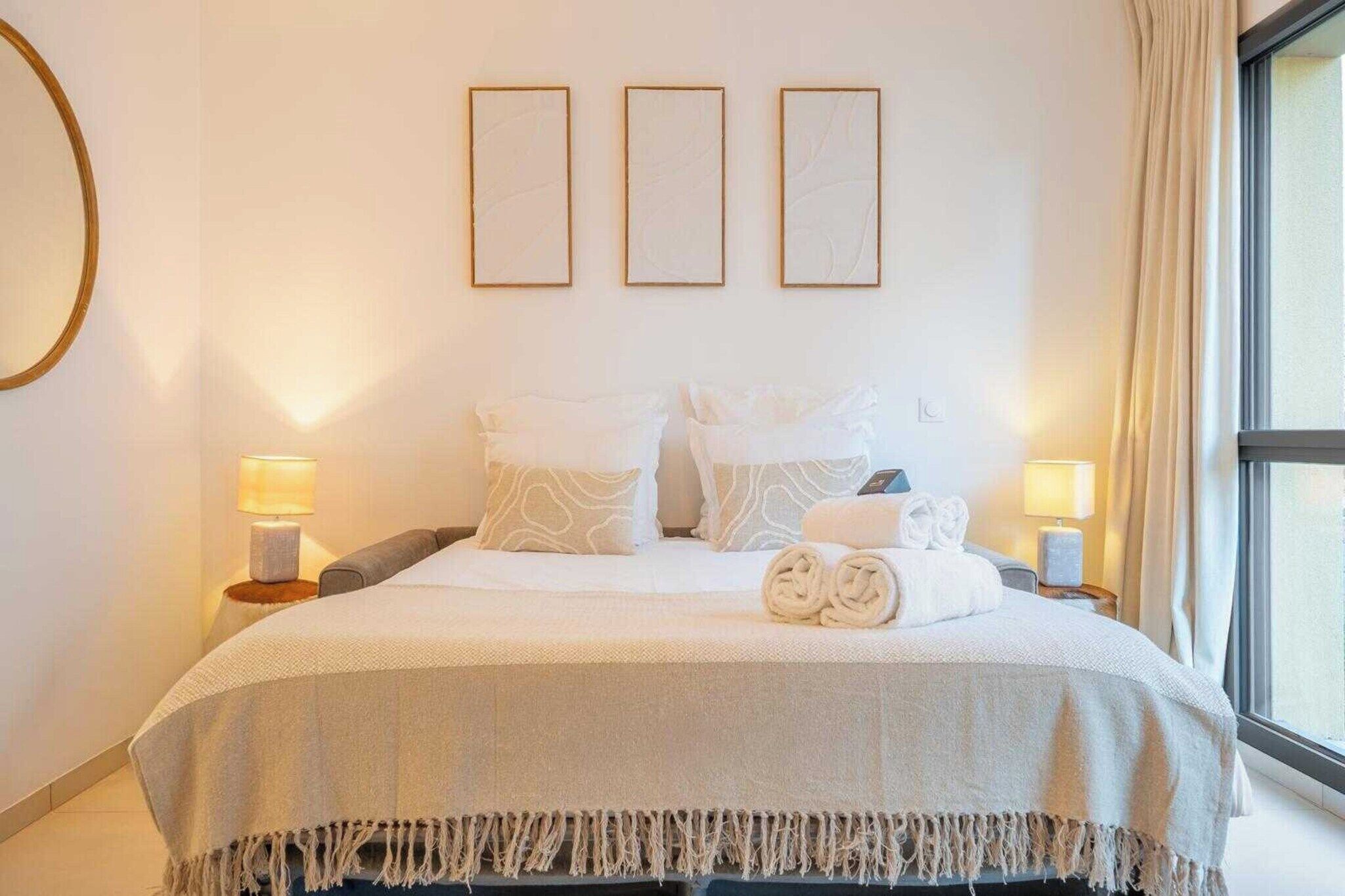 Una cama bien hecha con una colcha blanca y dos almohadas, flanqueada por lámparas de noche, en una habitación con un ventanal grande y tres cuadros en la pared.