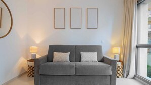 Un sofá gris con dos almohadones, flanqueado por dos mesitas de noche con lámparas, frente a una pared con tres cuadros enmarcados.