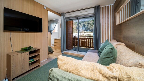 A modern room with a wooden TV stand, a sofa, and a sliding glass door leading to a balcony with a view of trees. - C. du Verdon N°3307 - 2P6 (Allos)