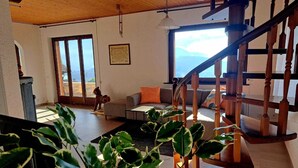 A spacious living room with a wooden staircase, a sofa, a coffee table, and a view of the outdoors through large windows. - Casa RuKi (Capriasca)