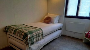 A small, single-bed room with a white bed, a green and white checkered blanket, a brown pillow, and a window with blinds. - Casa RuKi (Capriasca)