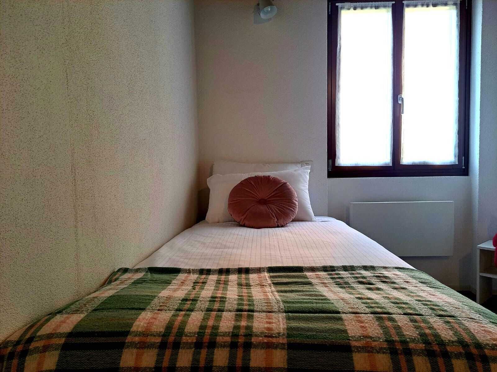 Een kleine eenpersoonskamer met een geruit sprei, een wit kussen en een raam met uitzicht.