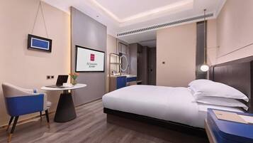 A modern hotel room with a bed, a desk, a chair, a TV, and a bathroom visible through an open door.