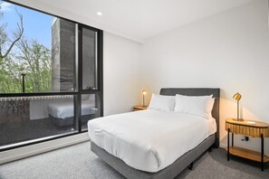 Ein modernes Schlafzimmer mit großem Fenster, einem Bett mit weißer Bettwäsche, zwei Nachttischen mit Lampen und Blick auf Bäume.