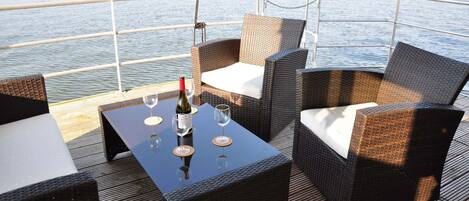 A deck with wicker furniture, a table with a bottle and glasses, and a view of the water.