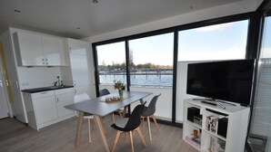 A modern kitchen with white cabinets, a dining area with a table and chairs, and a large TV on a white stand. - Hausboot Rosi mit Kajak in Ribnitz-Damgarten (Ribnitz-Damgarten)