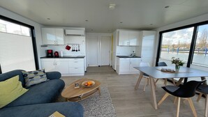 A modern interior with a sofa, coffee table, dining table, and chairs. There is a kitchen area with cabinets and a window overlooking a body of water. - Hausboot Rosi mit Kajak in Ribnitz-Damgarten (Ribnitz-Damgarten)