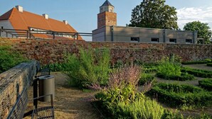 Um jardim com muro de tijolos, um banco de metal e um edifício com uma torre.