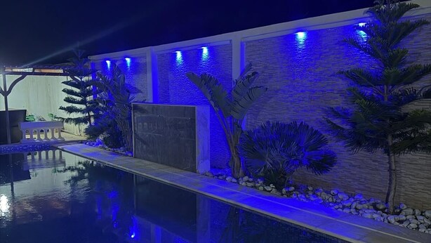 Een zwembad met betegeld zonnedek, een stenen muur met blauwe verlichting en tropische planten.