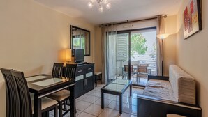 A modern living room with a glass dining table, black chairs, a sofa, a TV, and a ceiling light. - Carre Marine N°207 - 2P4 (Mandelieu-la-Napoule)