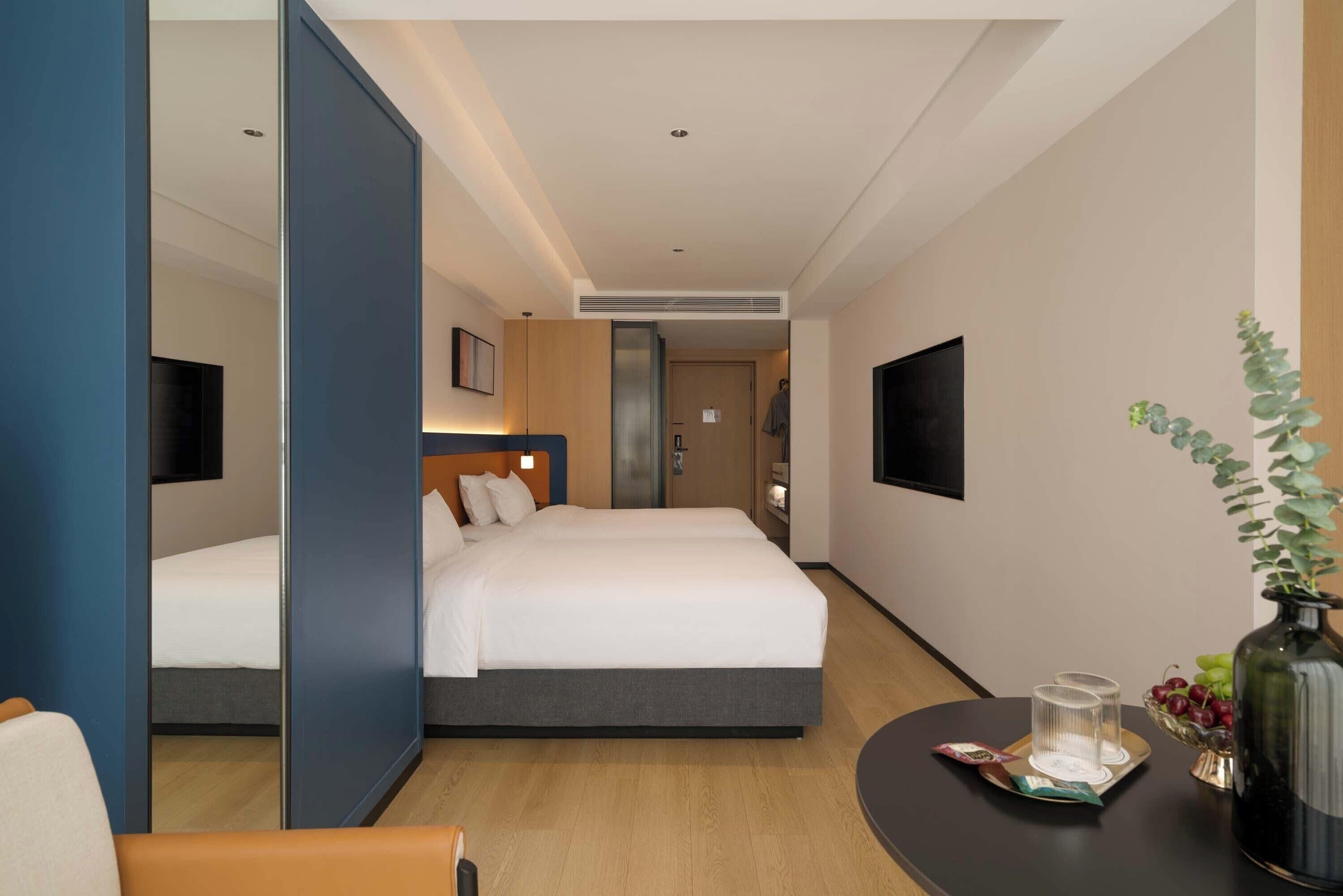 A hotel room with two beds, a TV, a bedside table with a vase and fruit, and a mirror.