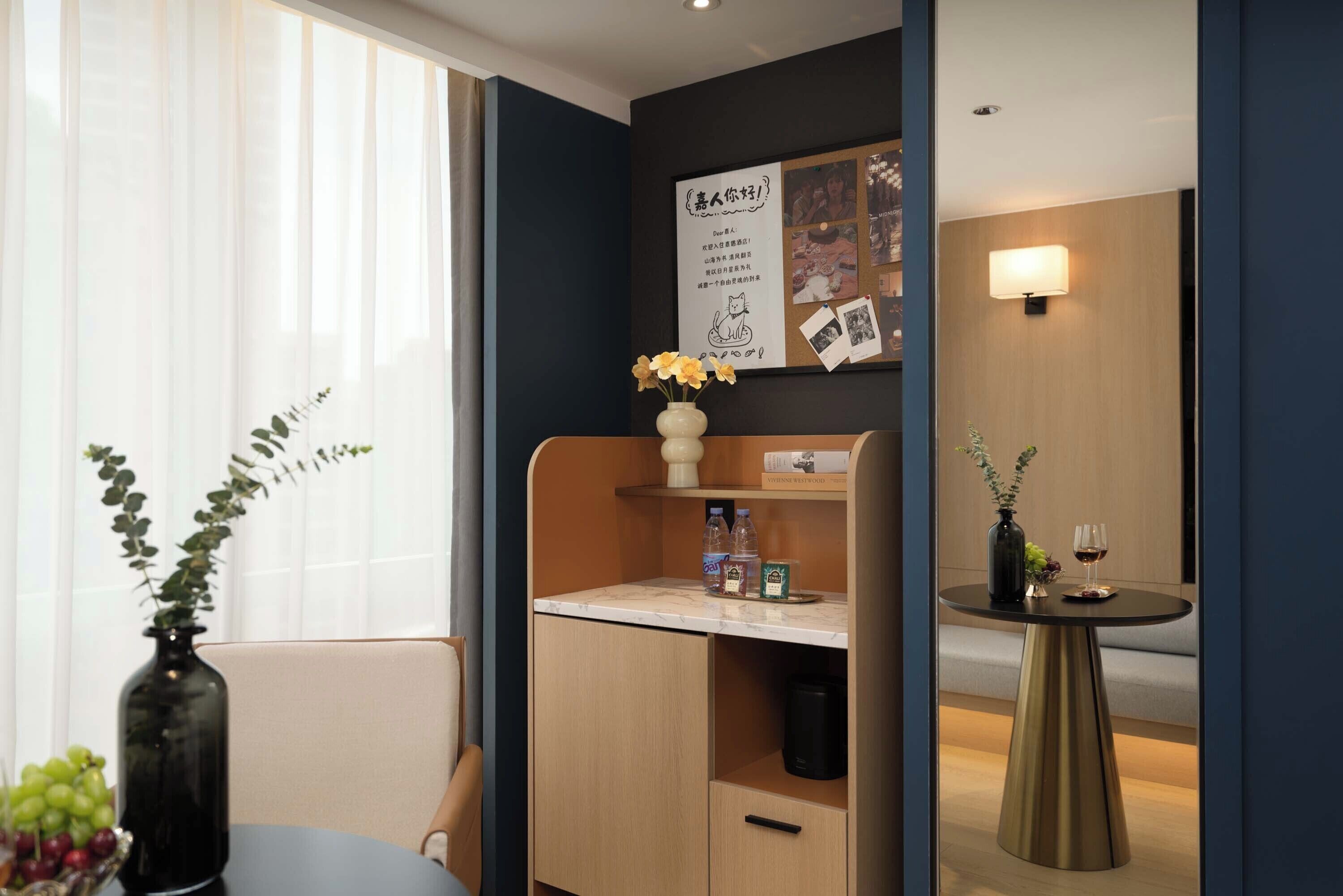 A hotel room with a wooden desk, a vase with flowers, a small table with a wine glass, and a bulletin board with photos and notes.