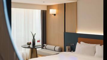 Een hotelkamer met een bed, een nachtkastje met een vaas en twee glaasjes, een zithoekje, en een bedieningspaneel aan de muur.