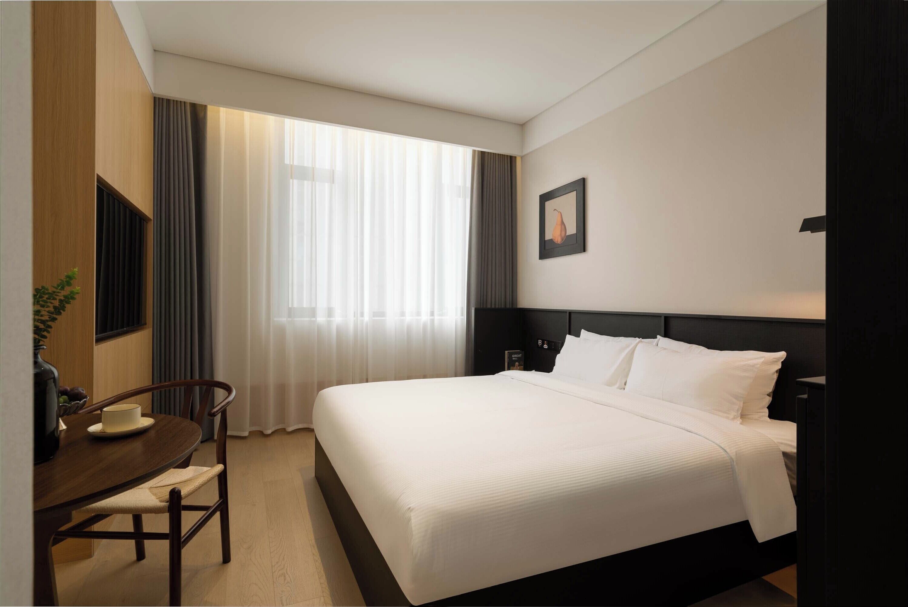 A hotel room with a large bed, a desk with a chair, a window with curtains, and a framed picture on the wall.