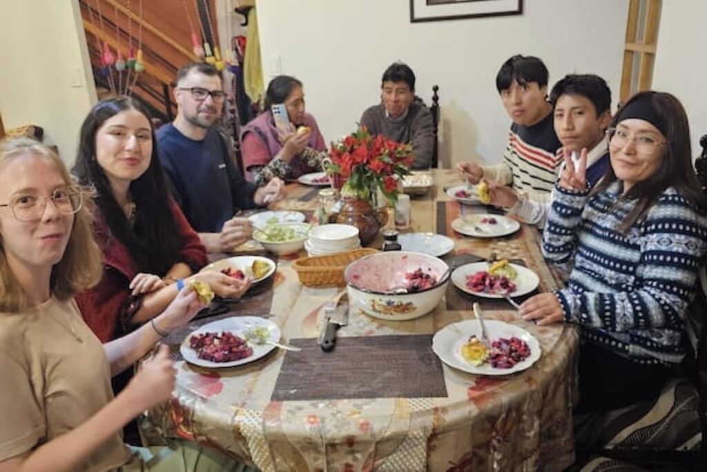 A group of people sitting around a table with various dishes, a vase with flowers, and a wooden table runner.