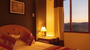 Une chambre d’hôtel avec un lit, une table de chevet, une lampe, un tableau encadré au mur et une fenêtre donnant sur le paysage.
