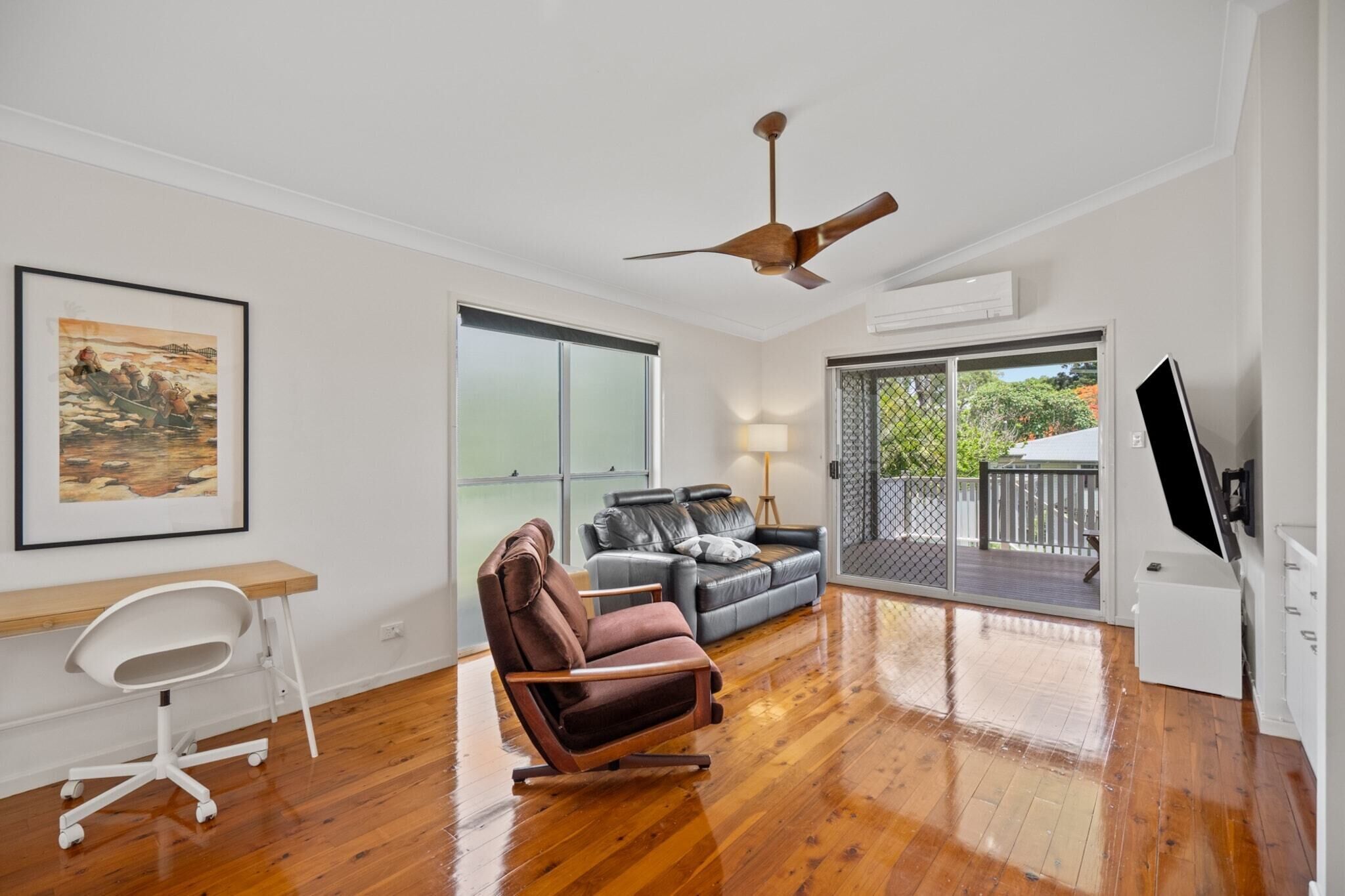 A spacious living room with a wooden floor, a ceiling fan, a large window, a sofa, a chair, and a TV.