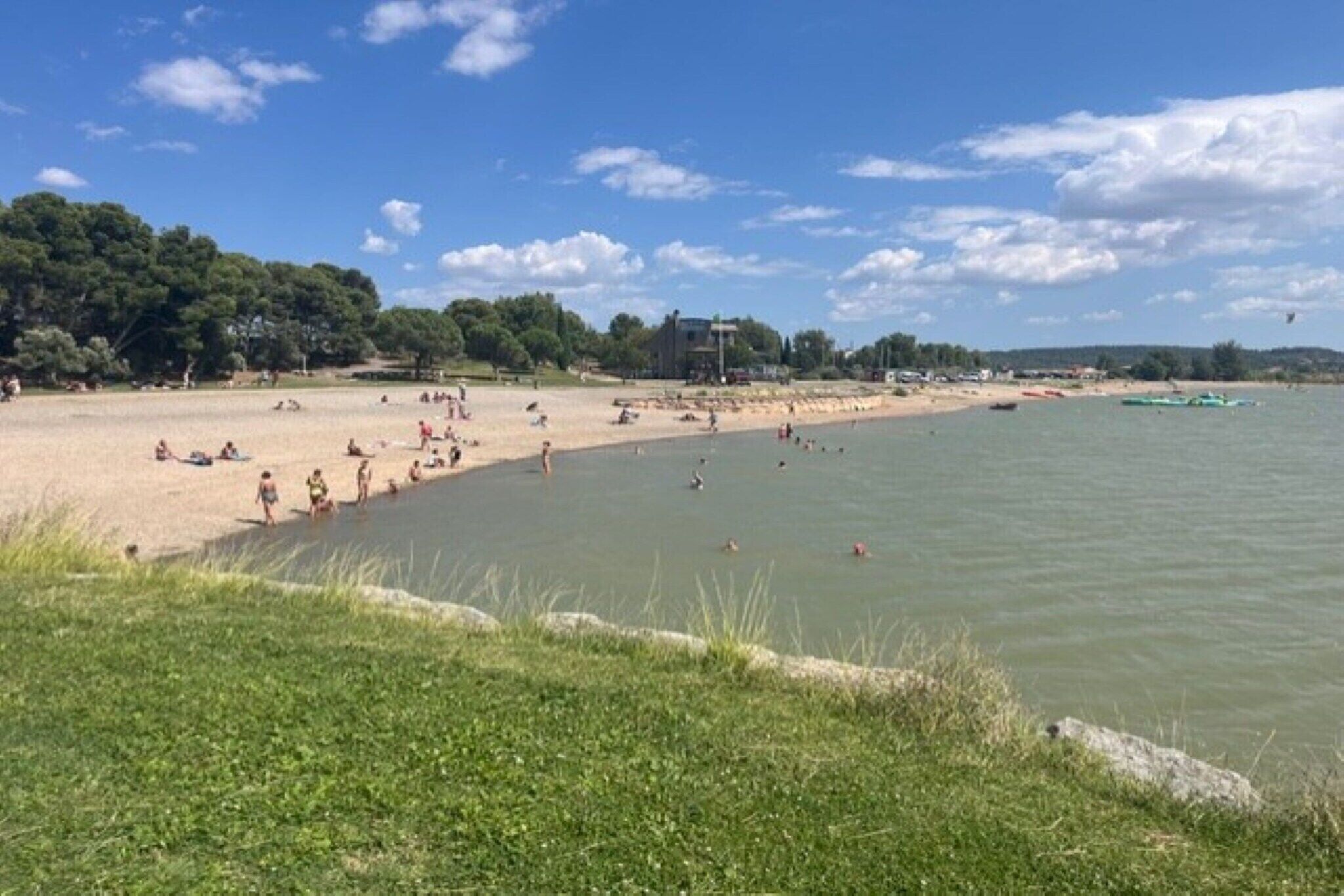 Een strand met mensen die zonnebaden en zwemmen, een grasveld op de voorgrond, en een gebouw op de achtergrond.