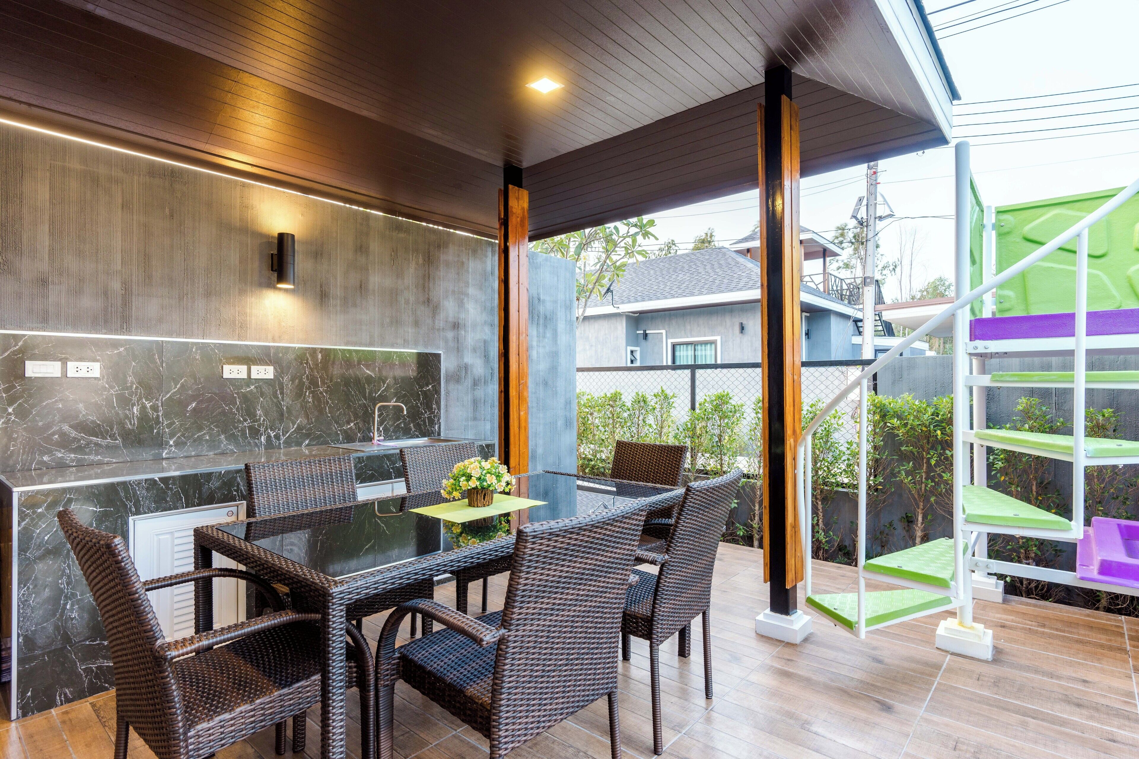 A modern outdoor dining area with a glass table and wicker chairs, a marble backsplash, and a view of a residential area.