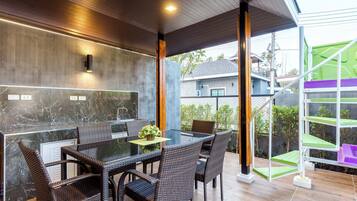 A modern outdoor dining area with a glass table and wicker chairs, a marble backsplash, and a view of a residential area.