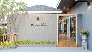 Una villa moderna con fachada de concreto, una escalera de madera que lleva a la entrada y una planta con flores a la derecha.