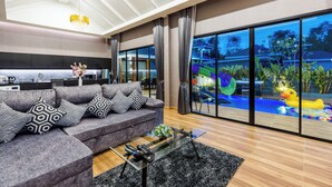 Una sala de estar moderna con un sofá gris, cojines estampados en blanco y negro, una mesa de centro de vidrio y vistas a una piscina y jardín a través de grandes puertas de cristal.