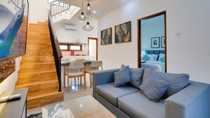 A modern living room with a grey sofa, a glass coffee table, and a wooden staircase. - Villa Sandalwood - Seminyak (Seminyak)