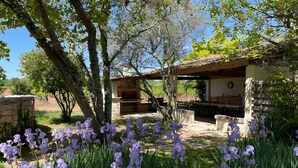A stone building with a brick oven, surrounded by trees and purple flowers.