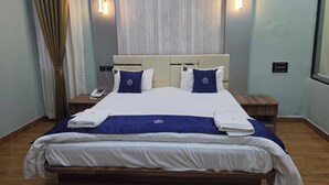 Una cama bien hecha con una colcha azul y blanca, dos almohadas y una manta a juego. Hay dos mesitas de noche con lámparas, un teléfono y un pequeño tocador de madera.