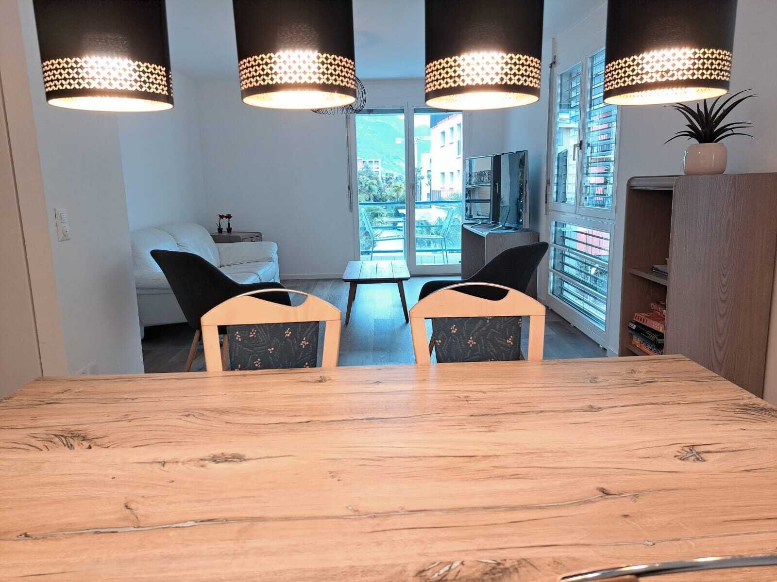 A dining area with a wooden table and two chairs, a sofa, and a TV in the background.