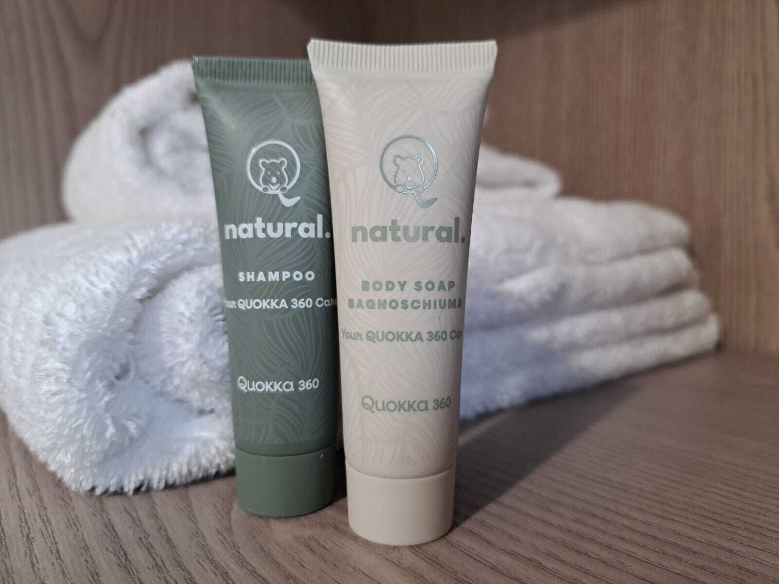 Deux bouteilles de shampooing et de savon pour le corps Quokka 360, à base d’ingrédients naturels, sont posées sur une surface en bois, avec une serviette blanche pliée en arrière-plan.