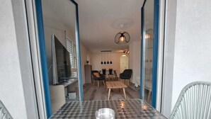 A modern dining area with a glass table, metal candle holder, and a view into a living space with a dining table, chairs, and a hanging light fixture. - Luisiana (Minusio)