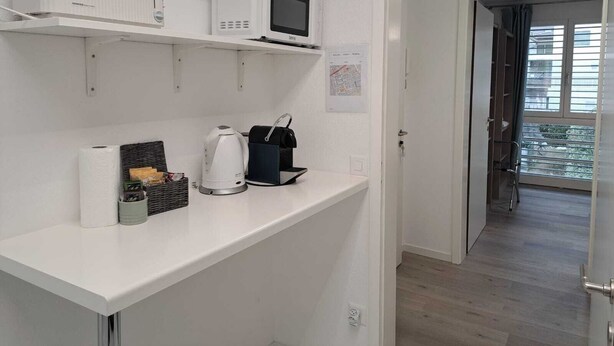 A modern kitchen with white cabinetry, a microwave, a kettle, and a coffee machine. There is a map on the wall and a door leading to a balcony. - Luisiana (Minusio)