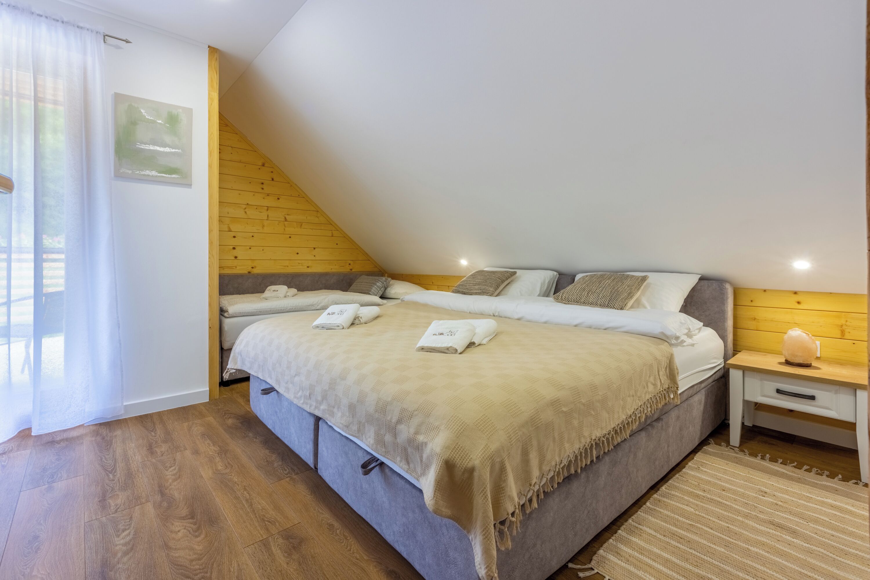 A bedroom with a sloped ceiling, a bed with a beige blanket, two bedside tables, and a window with curtains.
