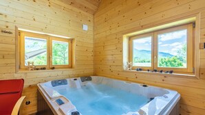 Une cabane en bois avec un bain à remous, deux fenêtres donnant sur les montagnes et un fauteuil rouge.
