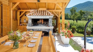 Un espace de restauration extérieur en bois, doté d’une cheminée en pierre, d’une longue table en bois dressée pour un repas, et offrant une vue panoramique sur un vignoble et des collines en arrière-plan.